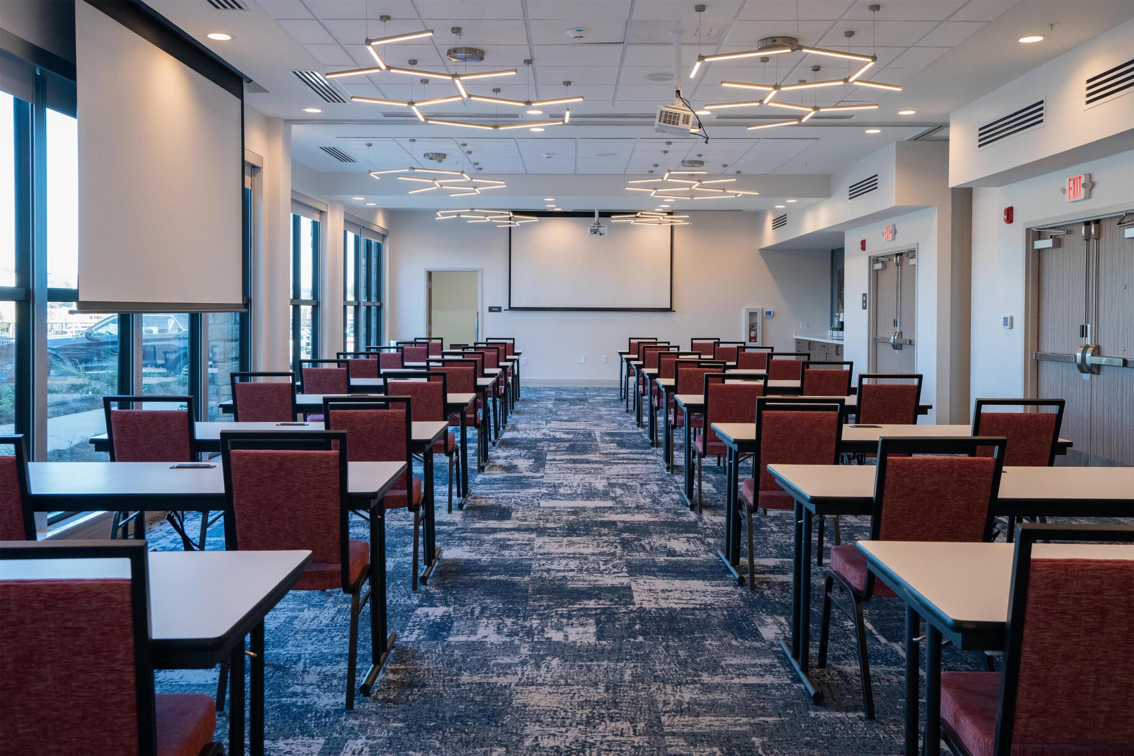 A conference room with rows of tables and chairs, large windows, and a carpeted floor.