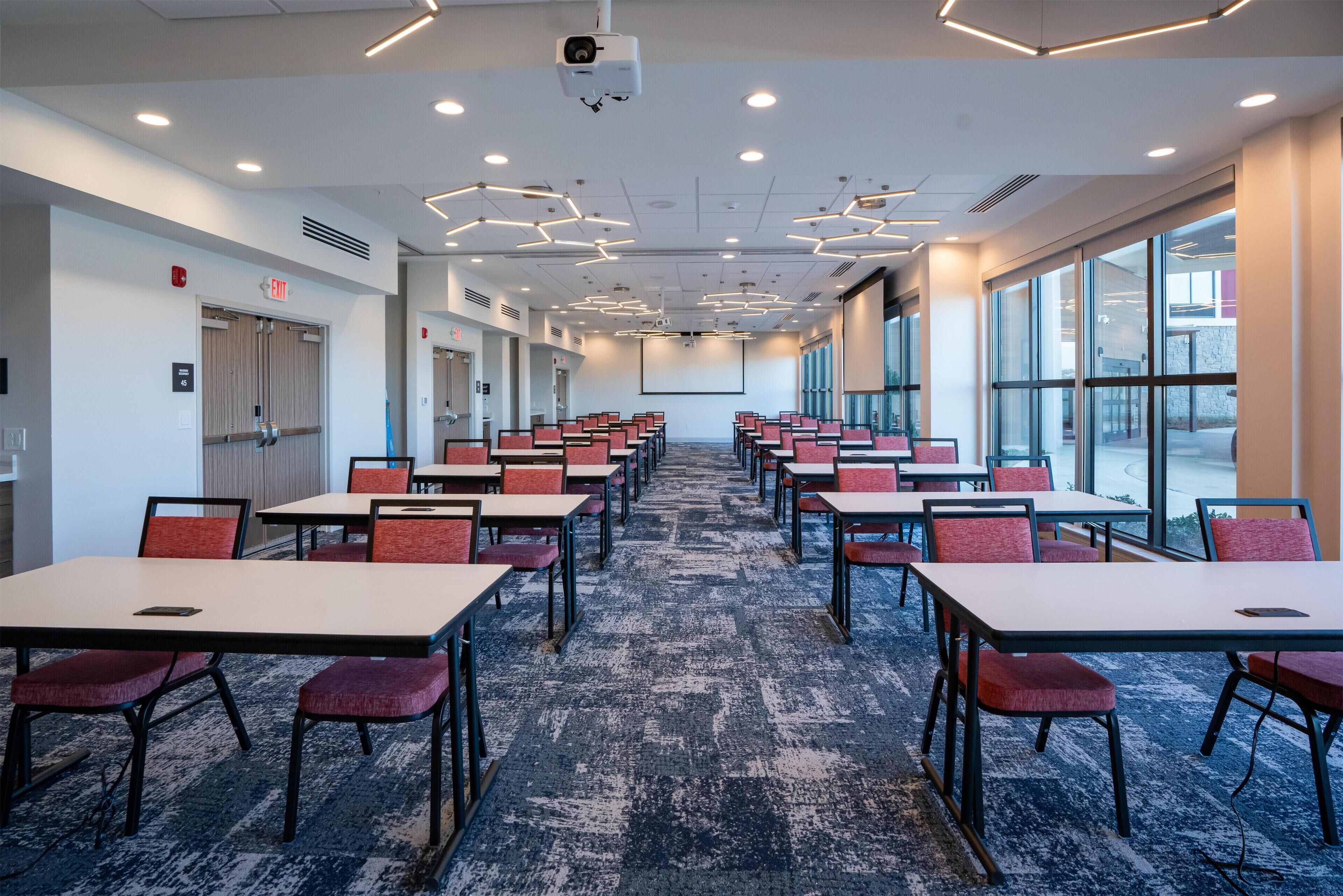A conference room with rows of tables and chairs, large windows, and a carpeted floor.