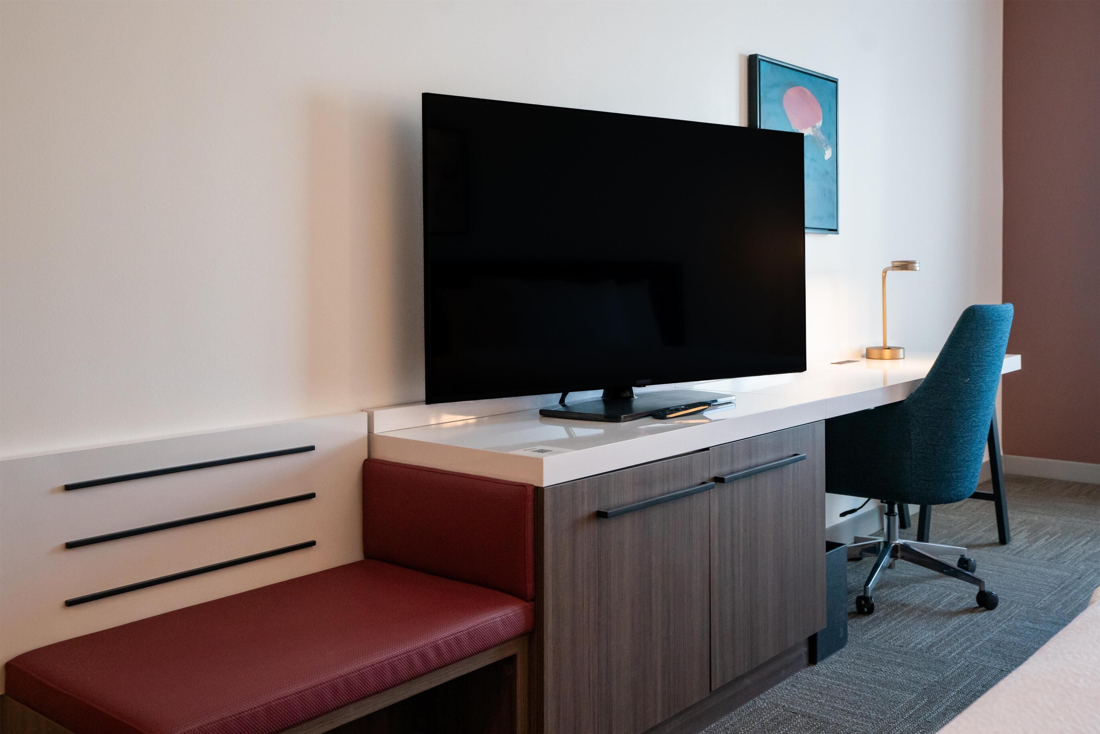 A modern hotel room with a flat-screen TV, a desk with a chair, a red bench, and a framed picture on the wall.