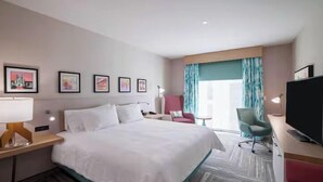 A hotel room with a large bed, a desk with a lamp, a TV, and a chair. There are framed pictures on the wall and a window with curtains. - Hilton Garden Inn Hoover Birmingham (Birmingham)