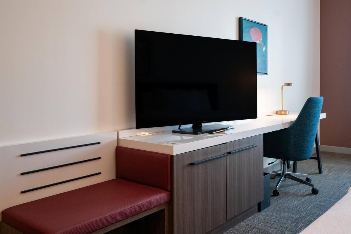 A modern hotel room with a flat-screen TV mounted on a white wall, a red cushioned bench, a blue office chair, and a desk with a lamp.