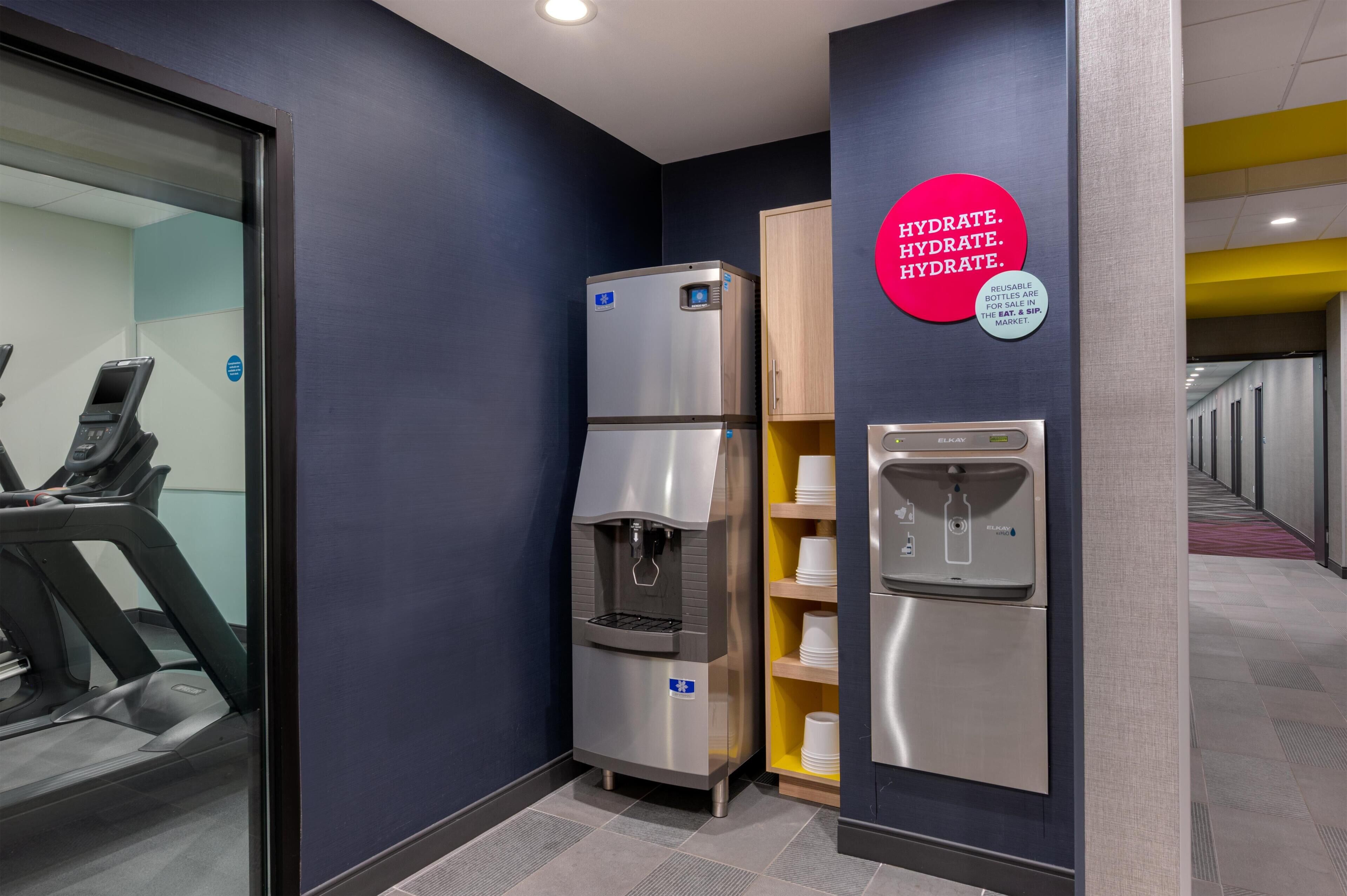 Ein Verpflegungsbereich mit Wasserspender und Kühlschrank, ein Laufband und eine Wand mit Aufkleber zur Trinkwahrheitsaufforderung.