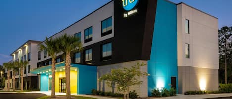 A modern hotel building with a prominent 'tru by Hilton' sign, featuring a mix of black and blue exterior panels, and a well-maintained lawn with palm trees.