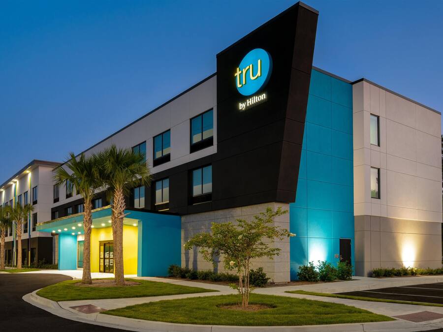 A modern hotel building with a prominent 'tru by Hilton' sign, featuring a mix of black and blue exterior panels, and a well-maintained lawn with palm trees.