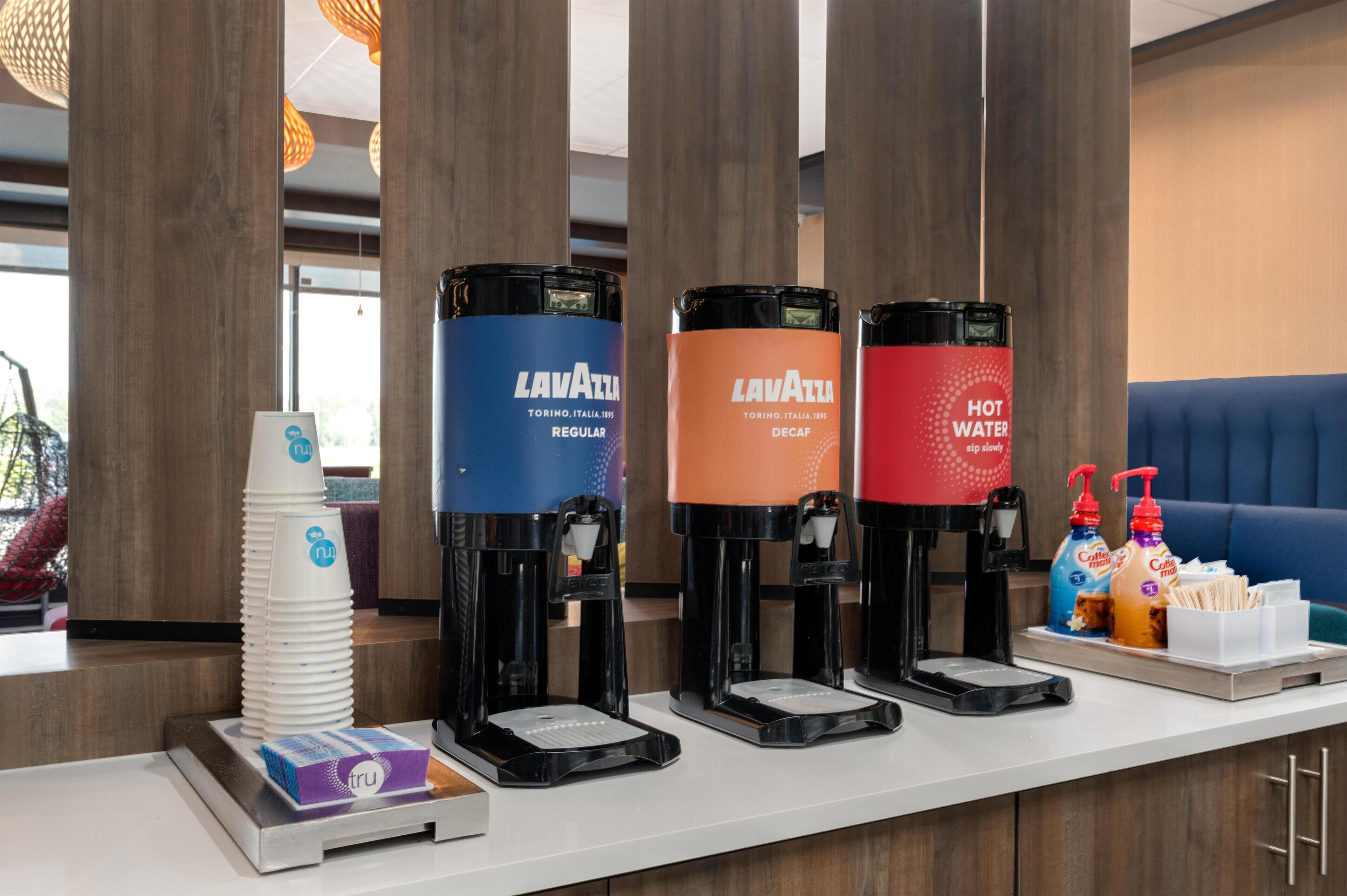 Un espace dédié au café, équipé de quatre distributeurs portant les inscriptions « Lavazza » et « EAU CHAUDE », situé dans un hôtel.