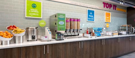 A breakfast bar with yogurt and oatmeal dispensers, a fruit bowl, and a sign for Top It Breakfast.