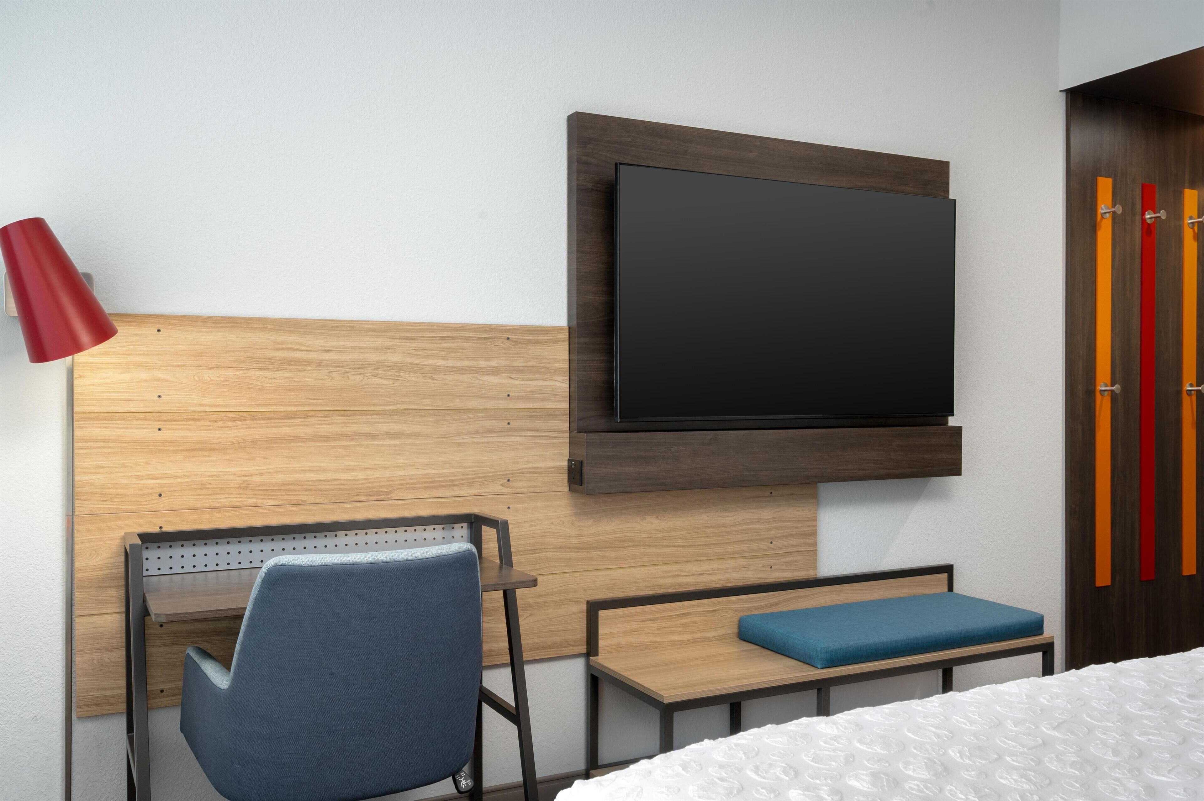A modern hotel room with a wooden headboard, a flat-screen TV mounted on the wall, a desk with a chair, a small table with a blue cushion, and a bed with white bedding.