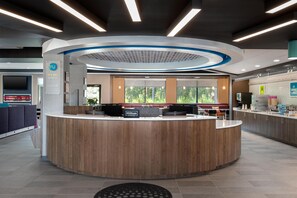 A modern hotel reception area with a curved reception desk, a digital screen, and a seating area with a view of the outdoors. - Tru By Hilton Brunswick (Brunswick)