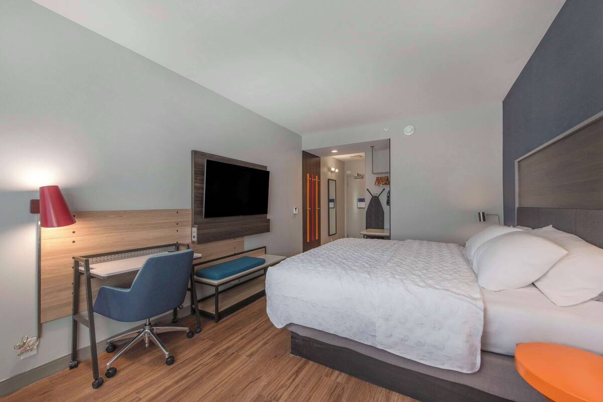 Habitación de hotel moderna con cama, escritorio, silla, televisor y un baño visible a través de una puerta abierta.