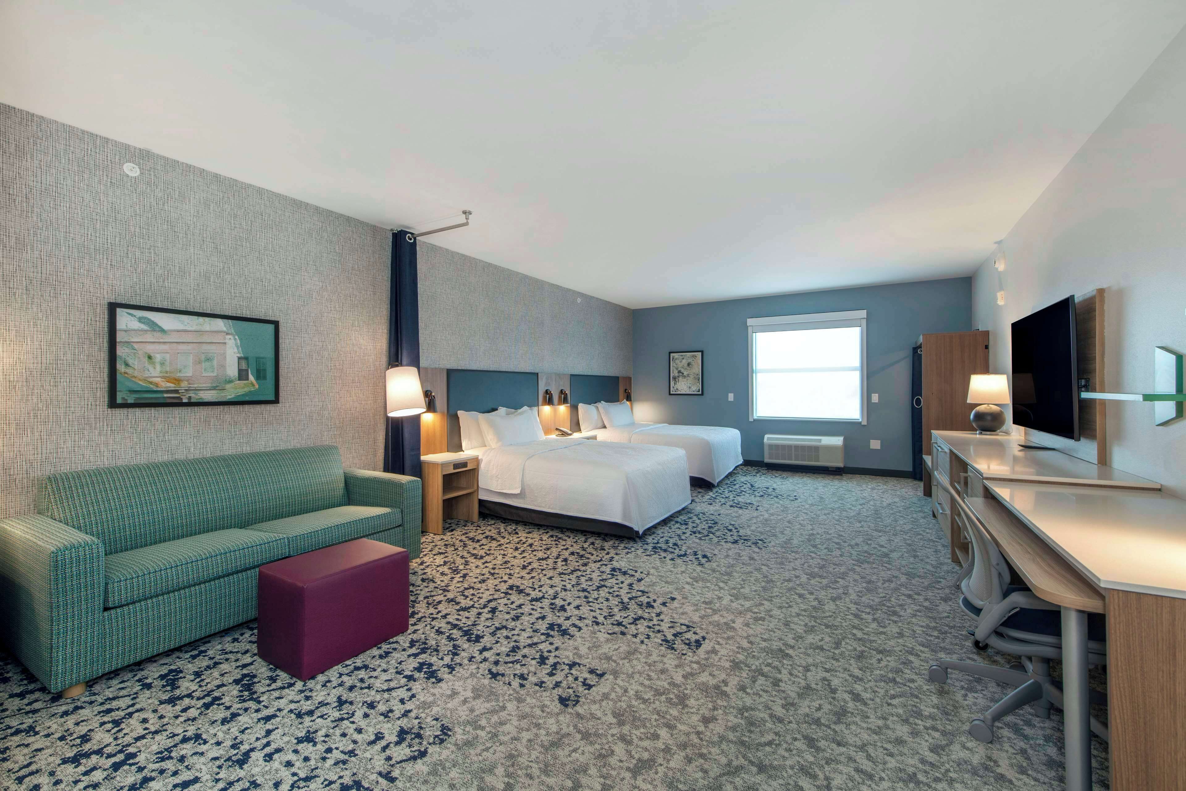 A hotel room with a green sofa, two beds, a desk, a TV, and a large window.