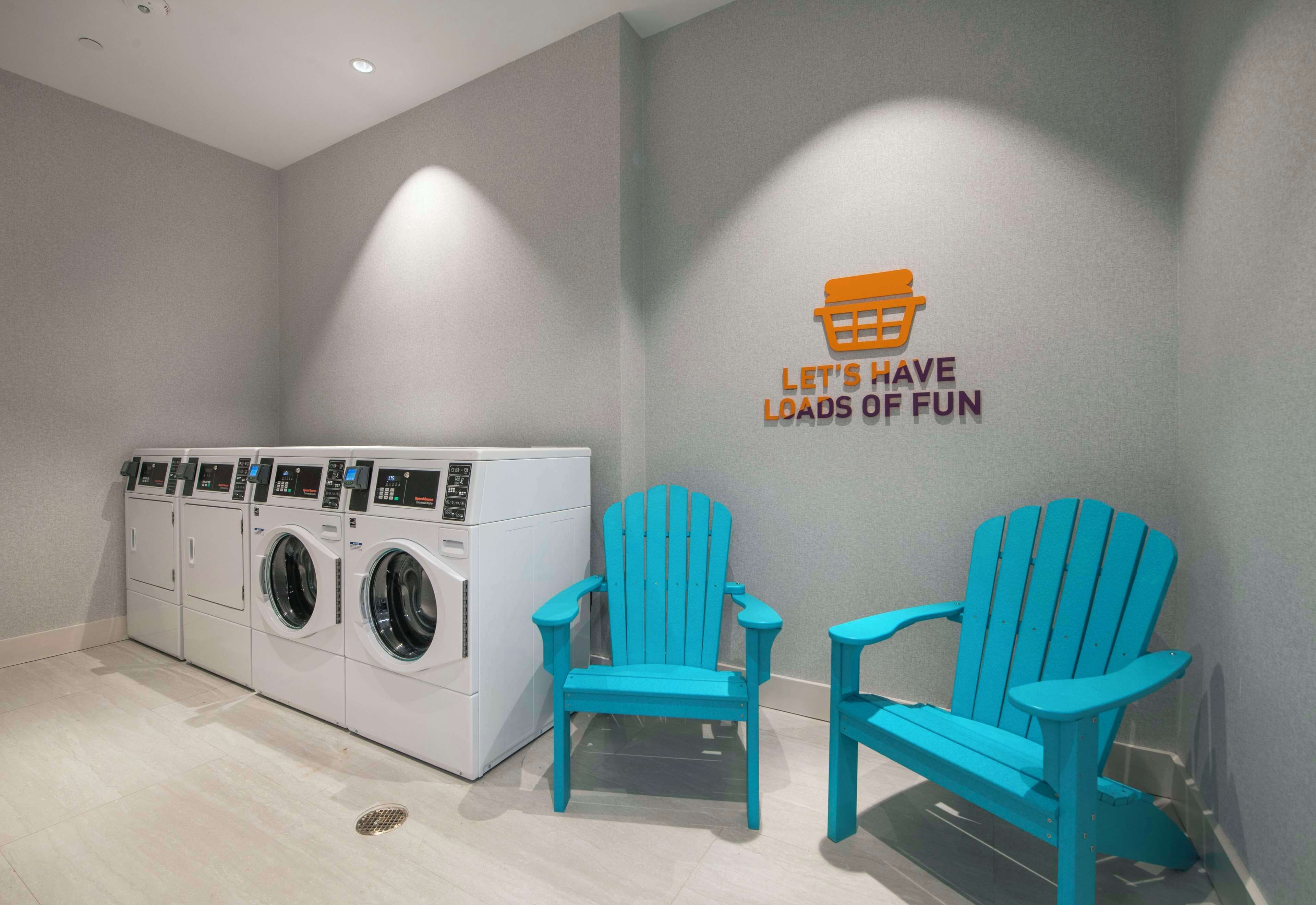A laundry area with a wall-mounted washer and dryer, two turquoise Adirondack chairs, and a wall sign that reads "LET'S HAVE LOADS OF FUN".