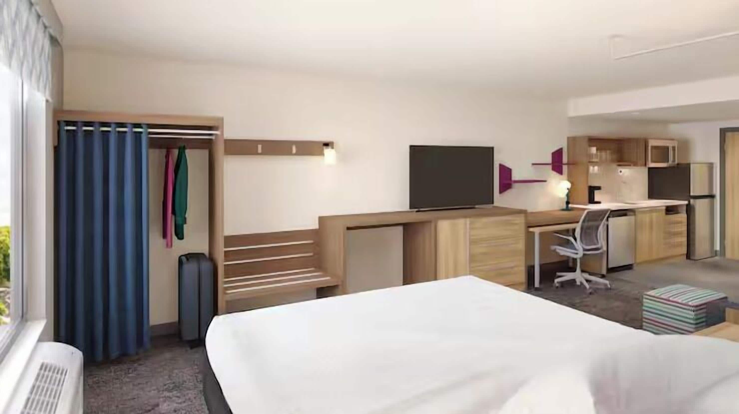 Habitación de hotel con cama, escritorio, televisor y kitchenette.
