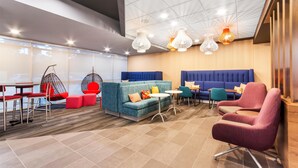A modern lounge area with seating arrangements, including a blue sofa, red chairs, and a pink armchair. There are tables and a hanging chair with red cushions. - Tru By Hilton Pensacola I-10 North Davis Highway (Pensacola)