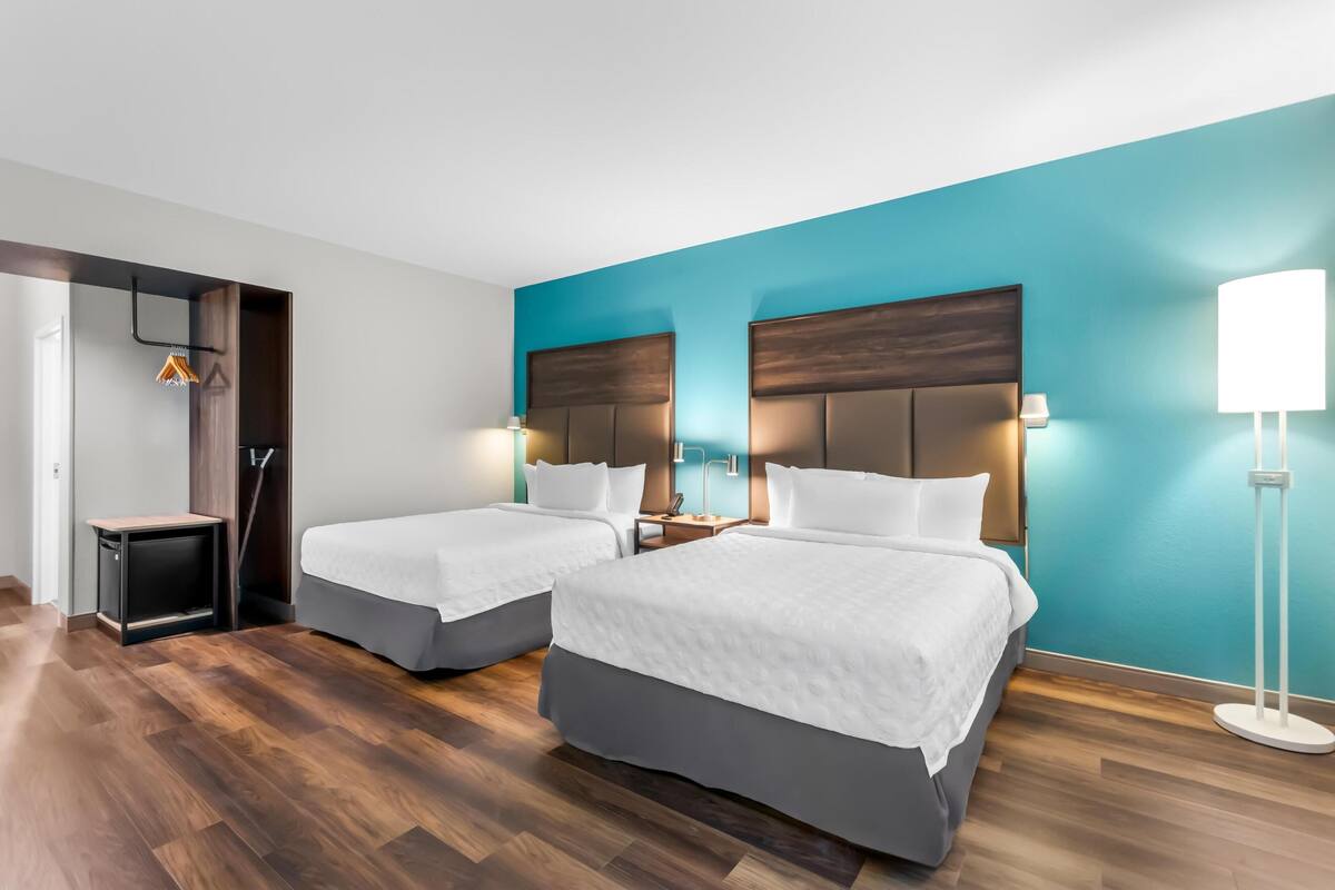 Quarto de hotel com duas camas, uma escrivaninha, uma cadeira, um abajur e um guarda-roupa.