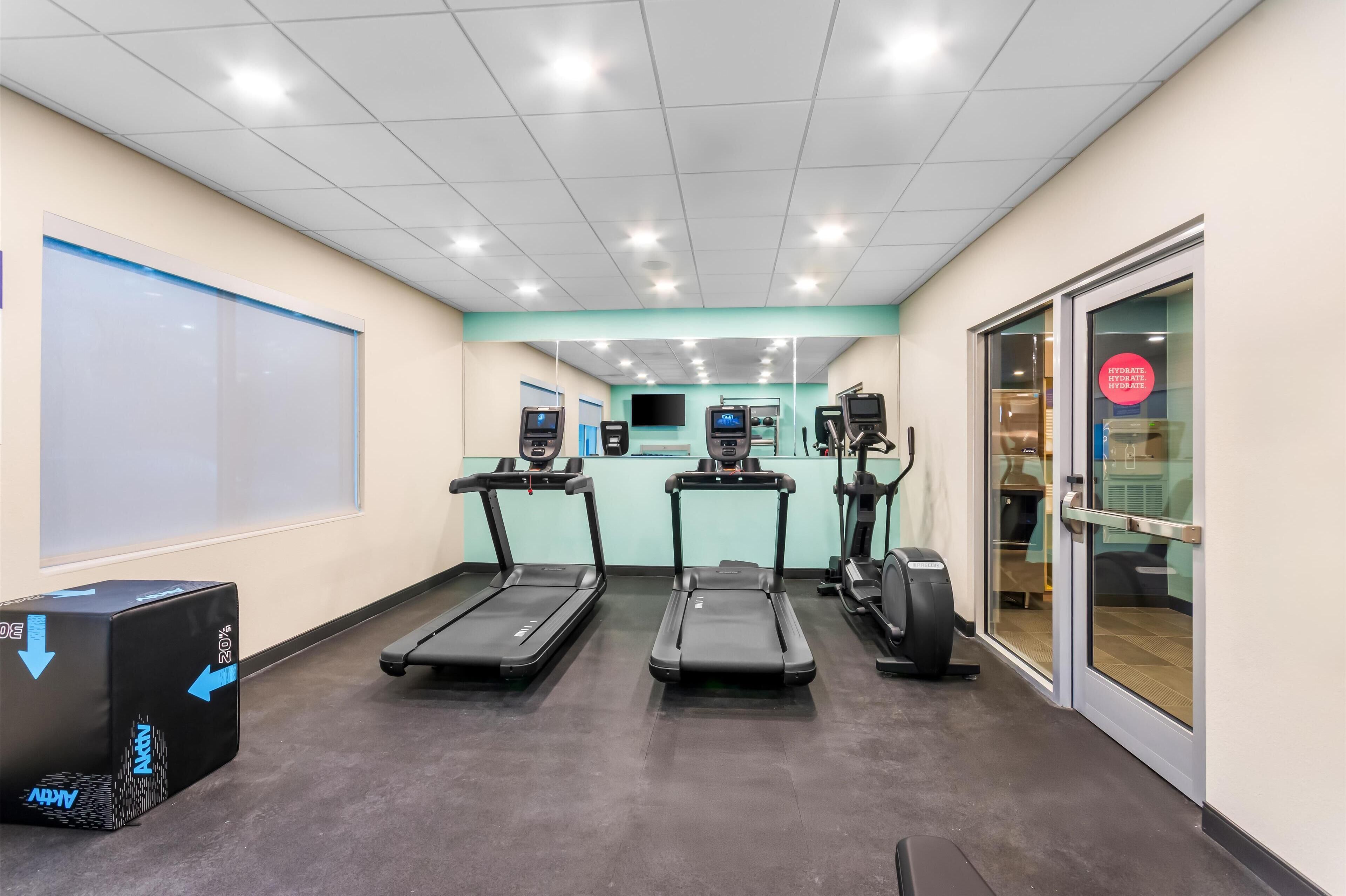 Ein Fitnessraum mit Laufbändern, Fahrrädern und einem Rudergerät. Es gibt Trainingsgeräte und eine Tafel an der Wand.