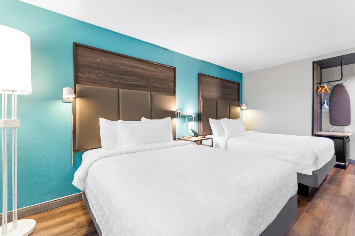 Quarto de hotel com duas camas, mesa de cabeceira, abajur e guarda-roupa.