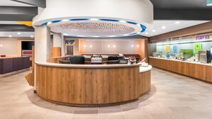 A modern hotel reception area with a wooden reception desk, a digital display, and a counter with amenities. - Tru By Hilton Pensacola I-10 North Davis Highway (Pensacola)