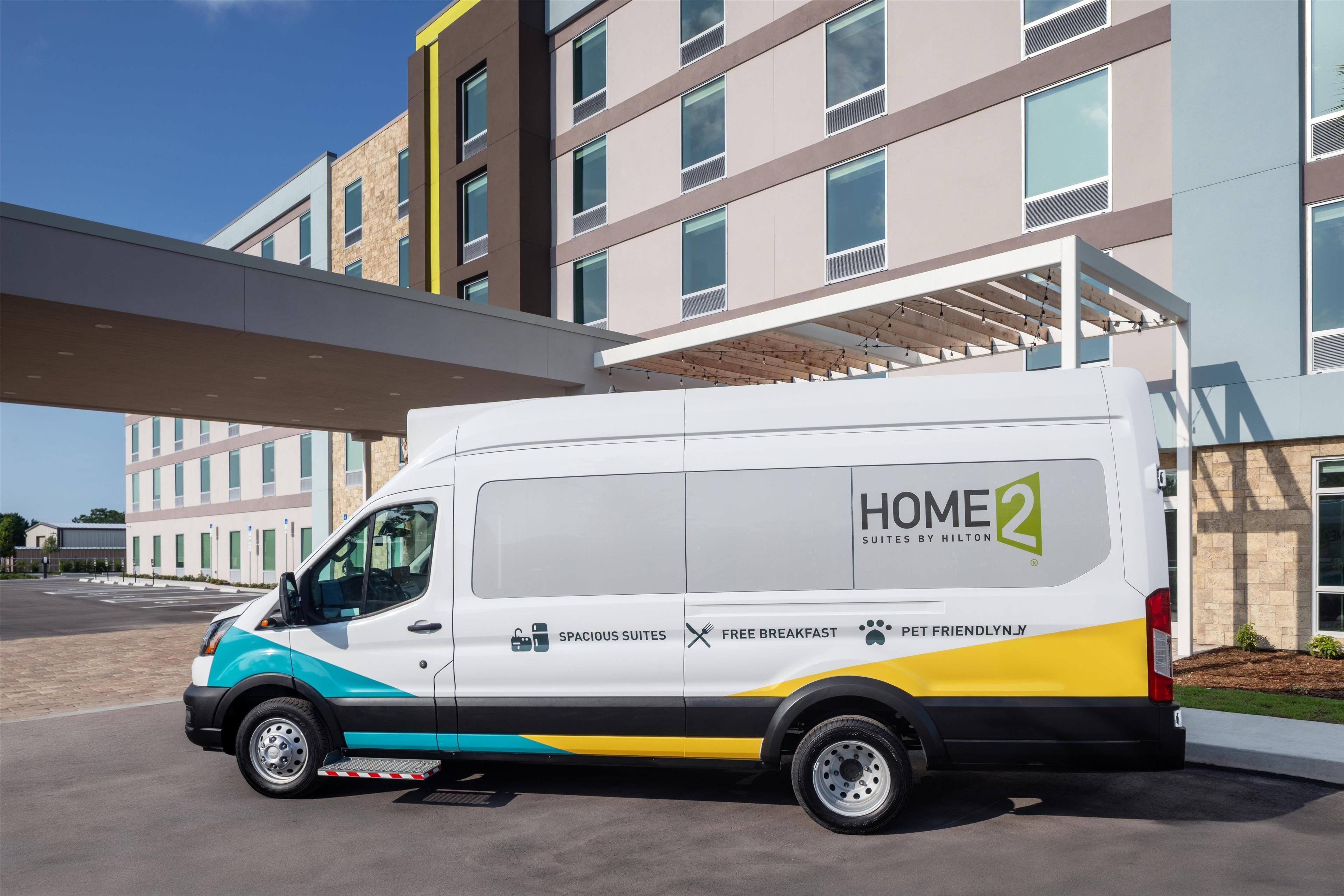 A white van with "HOME 2" and "SPACIOUS SUITES BY HILTON" branding parked in front of a modern building with multiple windows and a covered walkway.