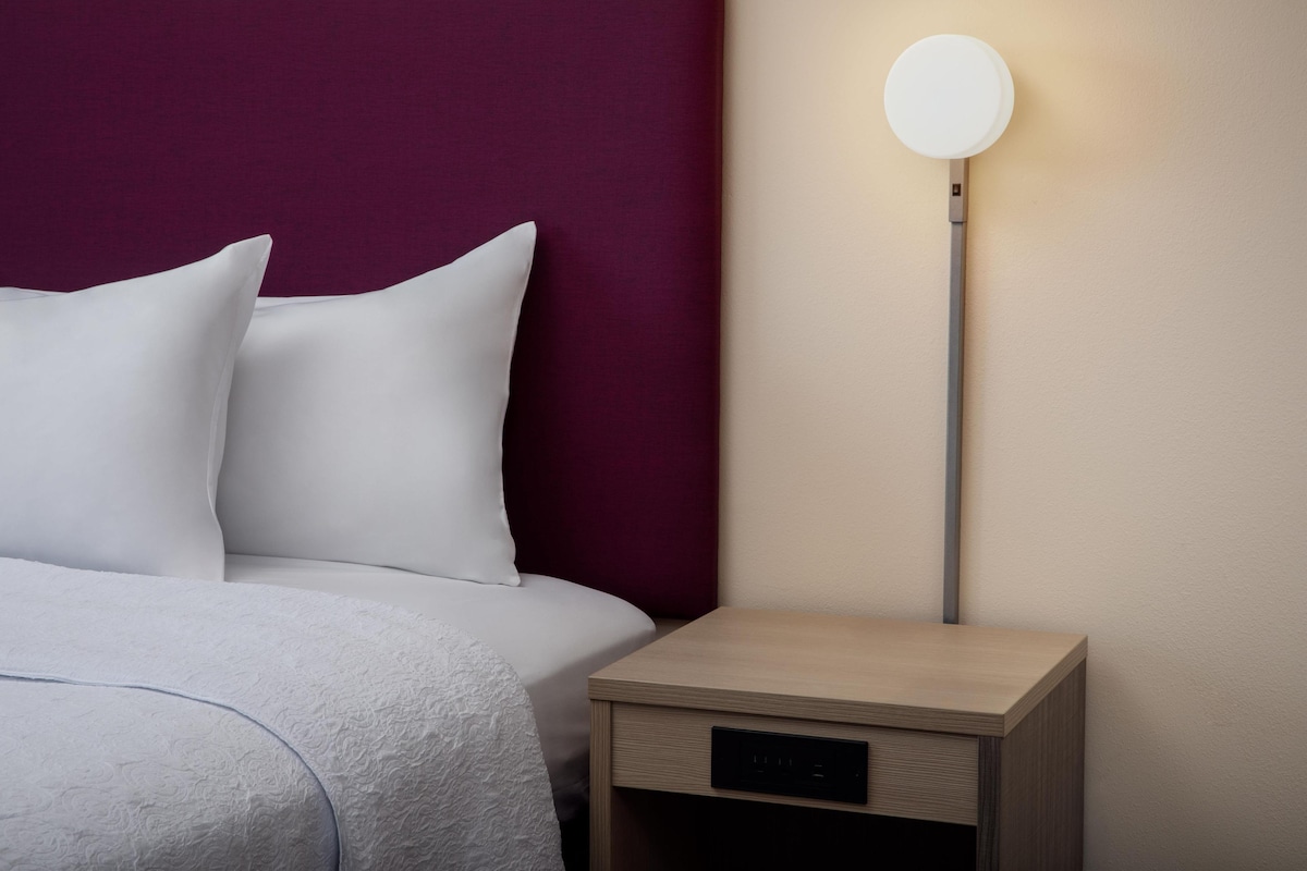 Un lit bien fait, avec du linge de lit blanc et des oreillers, une table de chevet avec une lampe, et un mur agrémenté d’une tête de lit de couleur foncée.