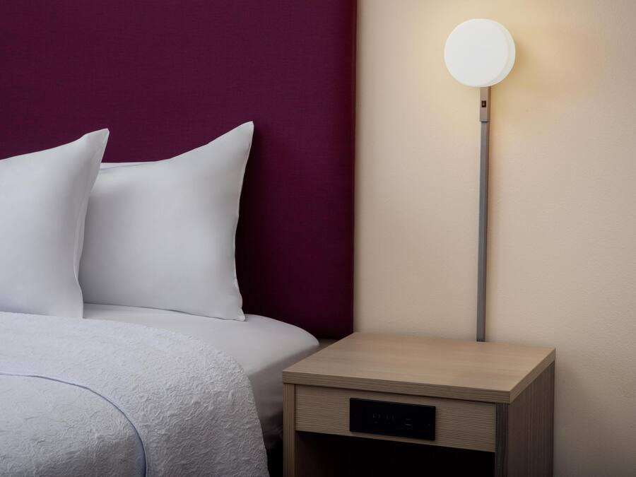 Una cama bien hecha con ropa blanca, una mesita de noche de madera con lámpara y un cabecero oscuro.
