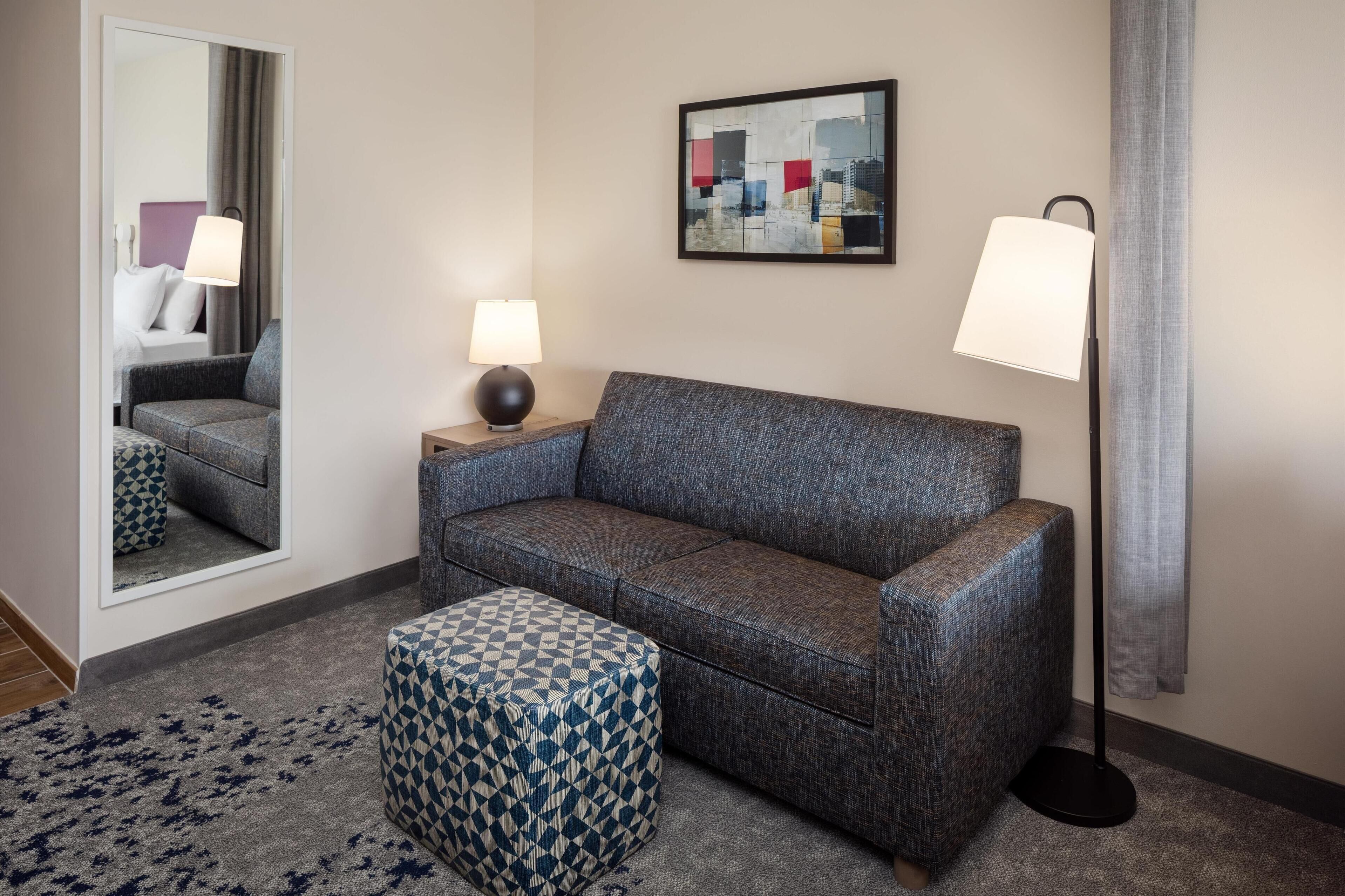 A modern hotel room with a grey sofa, a patterned ottoman, a bedside table with a lamp, a large mirror, and a framed cityscape artwork on the wall.