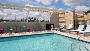 Zona de piscina con sillones de descanso y mesas, una pérgola y un muro de piedra.