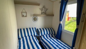 Een compacte kamer met twee bedden, voorzien van blauw-witte strepen in het beddengoed, een klein plankje met decoratieve spullen en een raam met gordijnen.