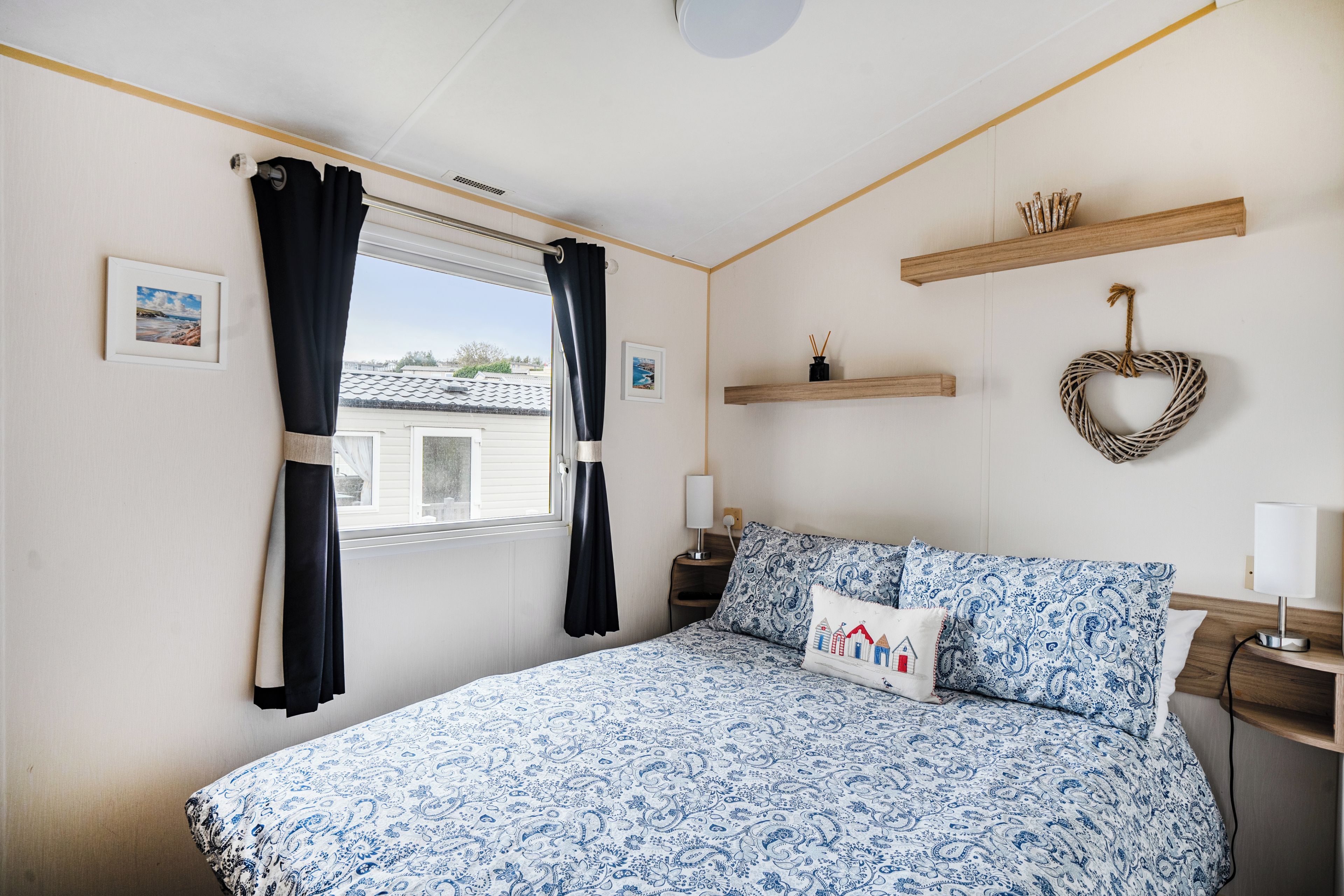 Une chambre à coucher avec un lit, des rideaux, une table de chevet, une lampe, une étagère fixée au mur et un cadre photo.