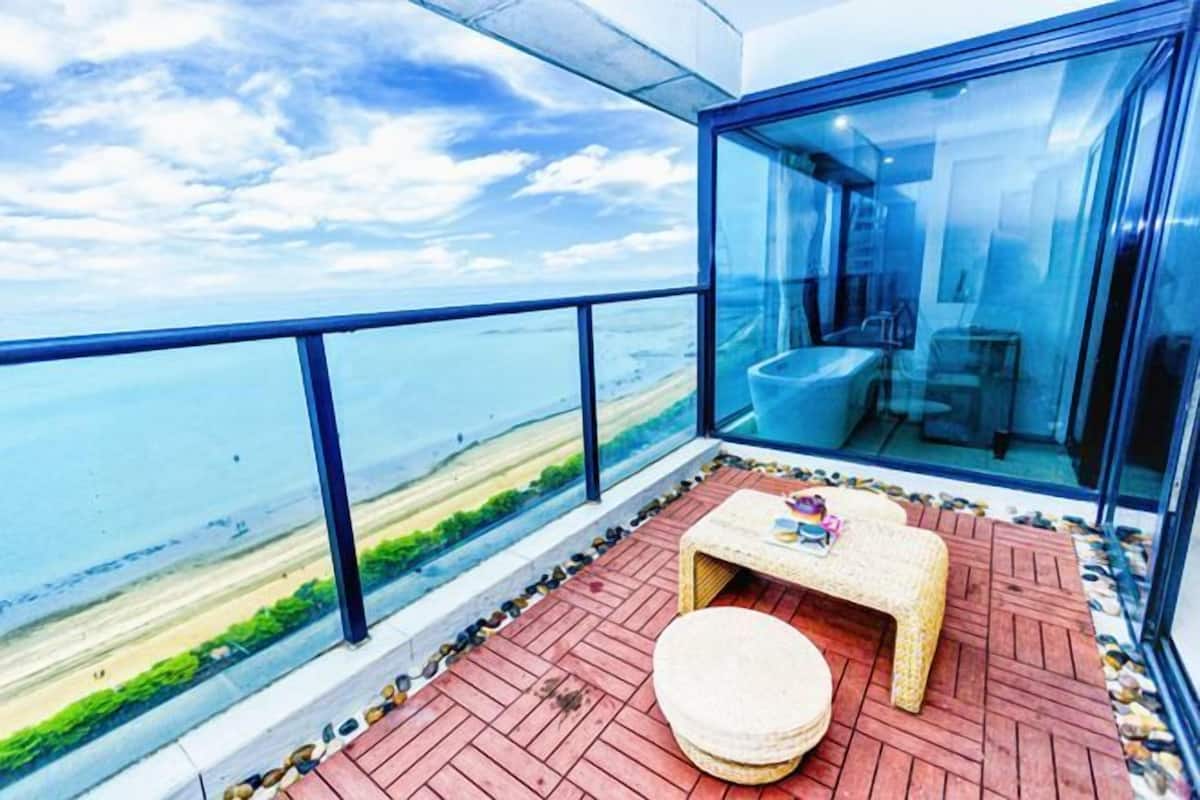 Ein Balkon mit Blick auf den Strand, eine kleine Sitzecke mit Tisch und Hocker sowie ein verglastes Badezimmer.