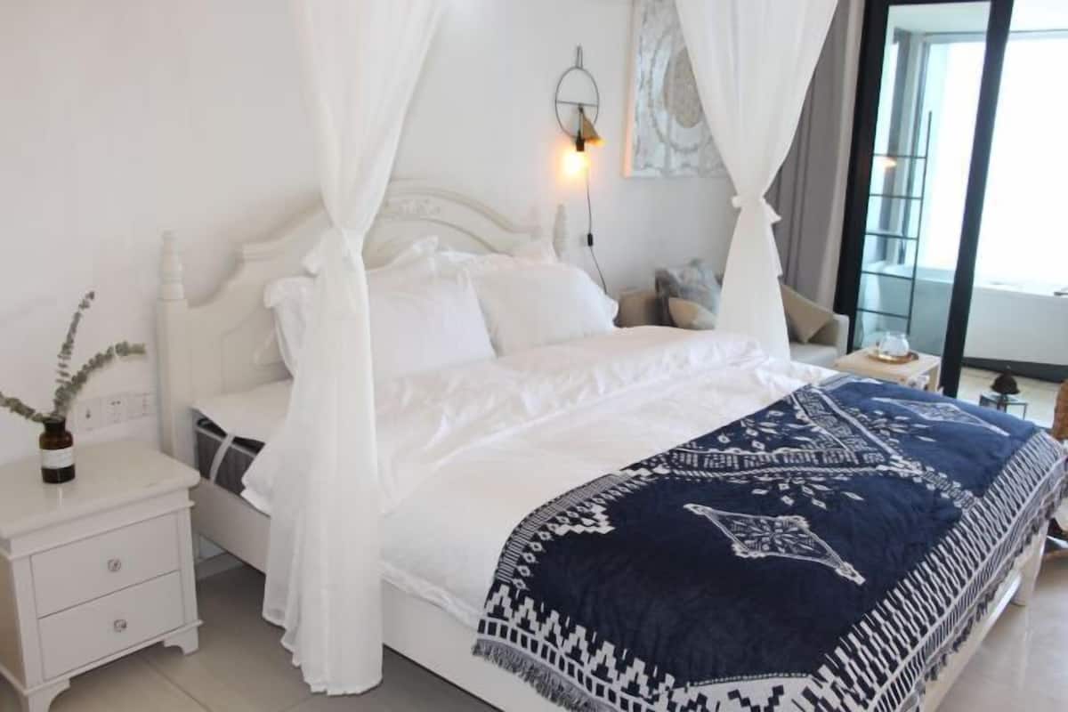 Ein Schlafzimmer mit einem Bett, einem weißen Nachttisch, einem Beistelltisch und einem Fenster mit Vorhängen.
