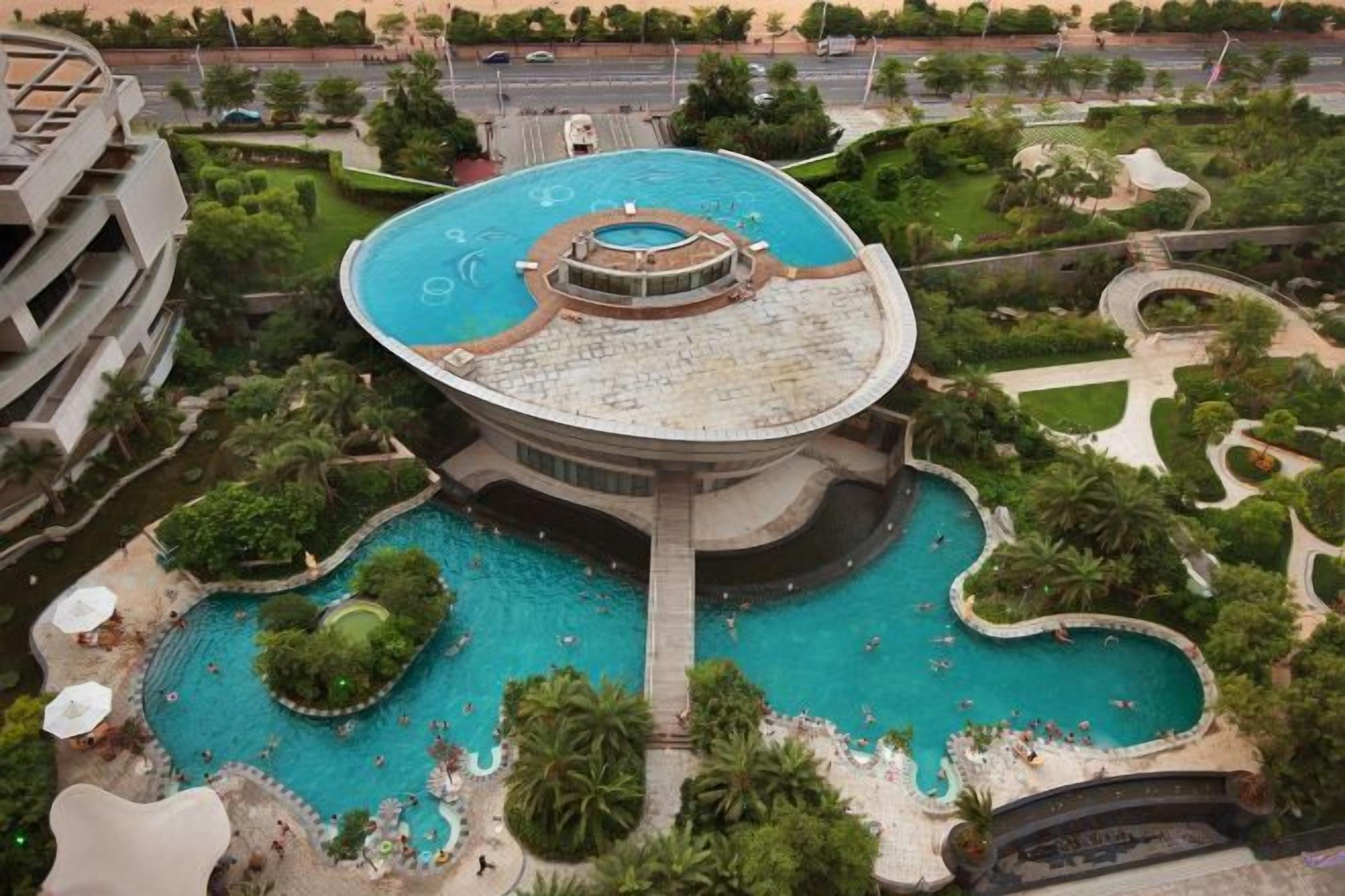 A large circular swimming pool with a central fountain, surrounded by lush greenery and palm trees.