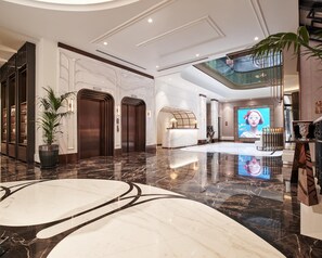 Un vestíbulo amplio con suelo de mármol, un mostrador de recepción, una gran pantalla digital y una escalera que conduce a un nivel superior.