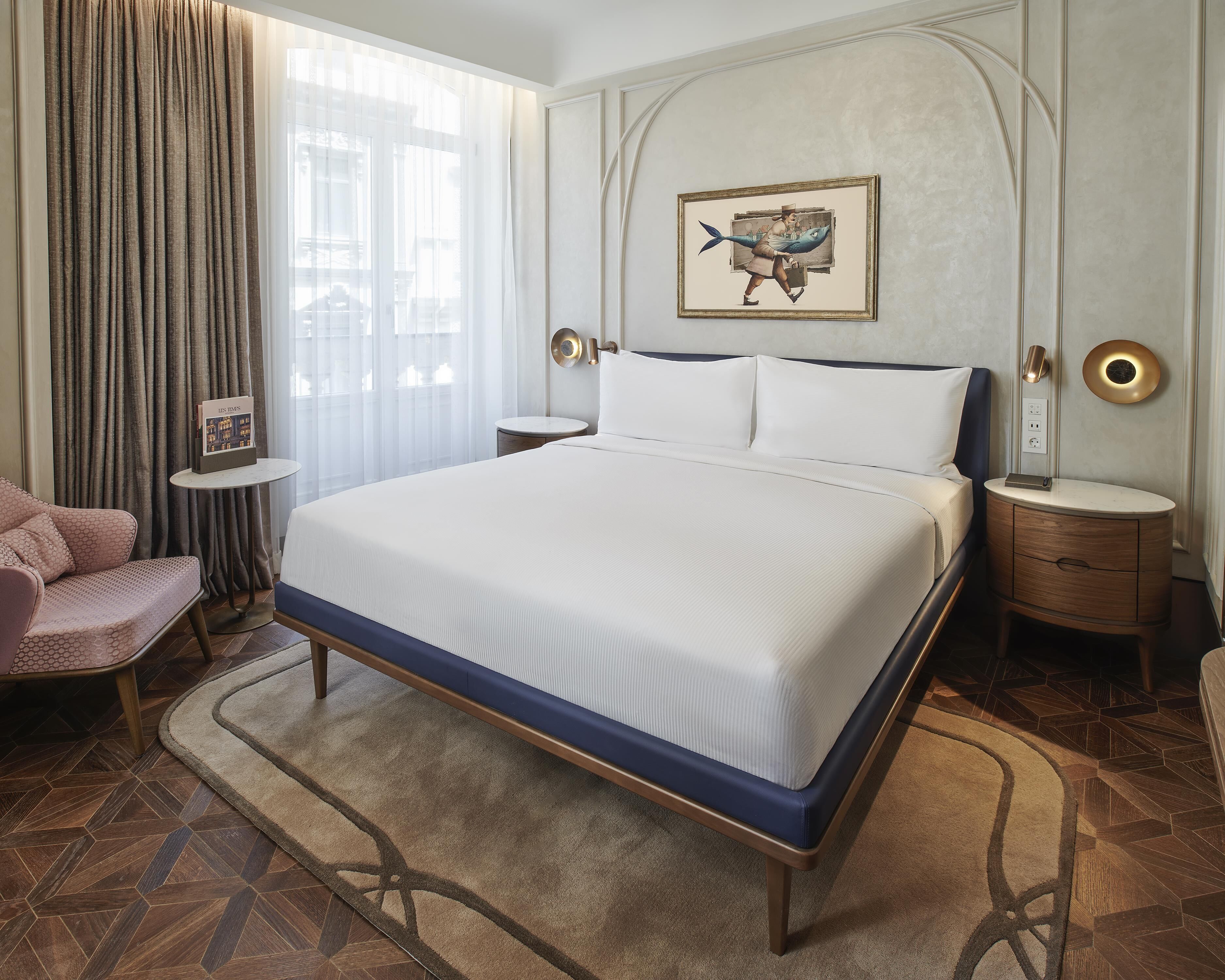 A hotel room with a large bed, a bedside table, a nightstand, a chair, a window with curtains, and a wall-mounted artwork.