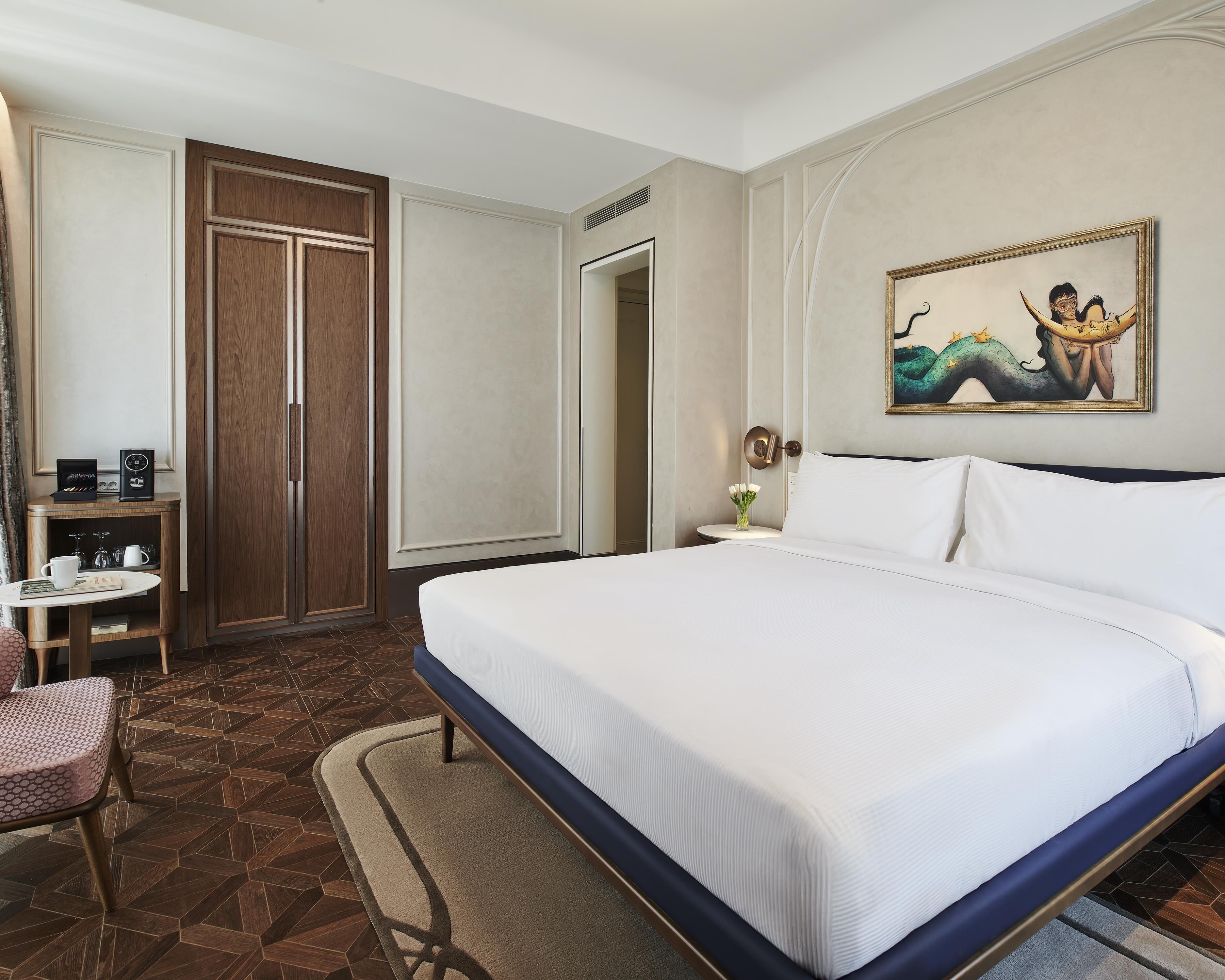 A hotel room with a bed, a desk, a chair, a wardrobe, and a painting on the wall.