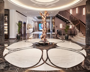A lobby with a marble floor, a central sculpture, a staircase, and a reception desk.