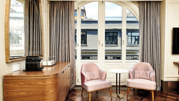 A hotel room with a wooden dresser, two pink chairs, a small table, and a large window with curtains. - Les Temps Istanbul Karakoy, Curio Collection By Hilton (Istanbul)
