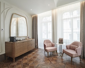 A hotel room with a wooden dresser, two pink armchairs, a small table, and a large mirror.