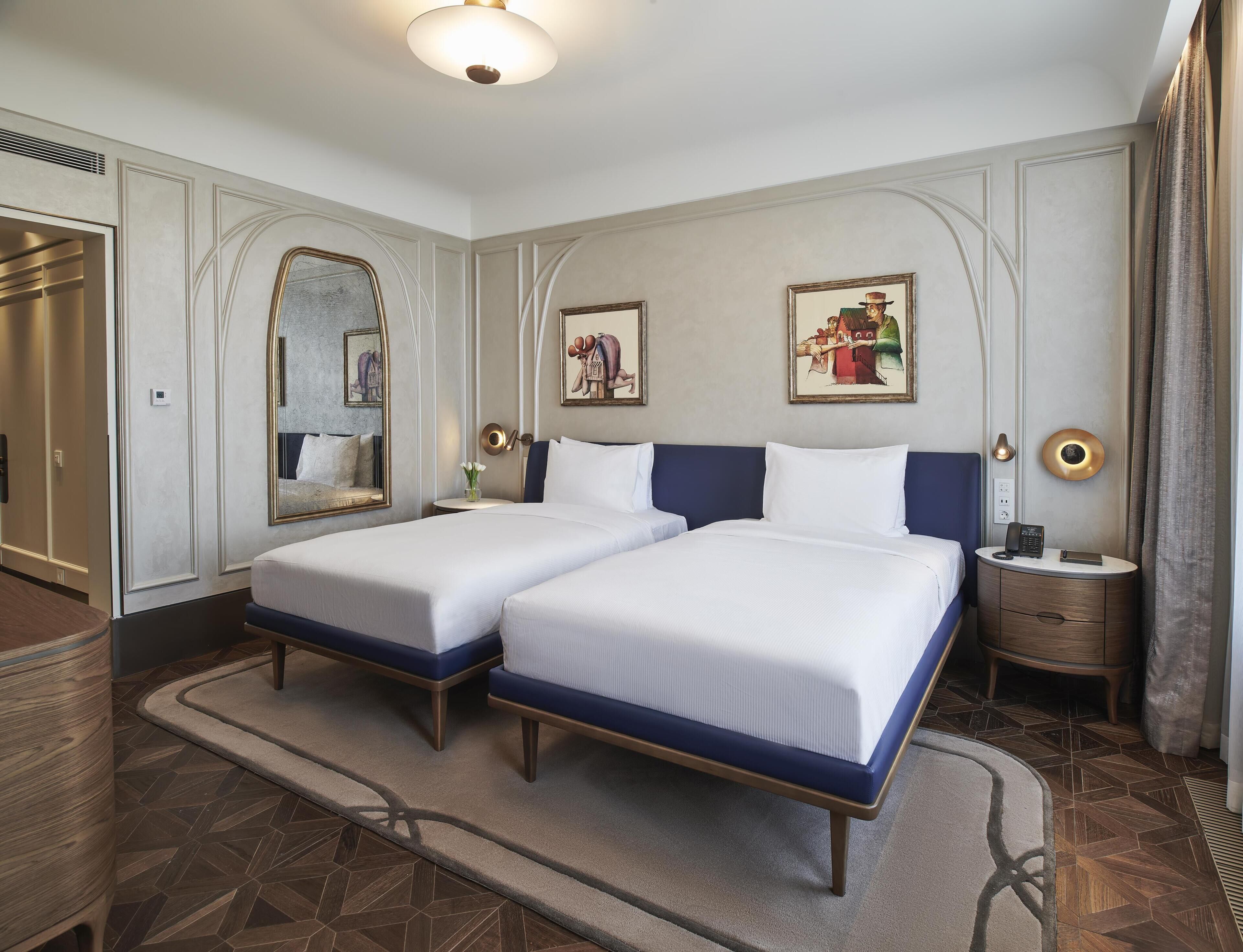 A hotel room with two beds, a mirror, a painting, a bedside table with a lamp, and a wardrobe.