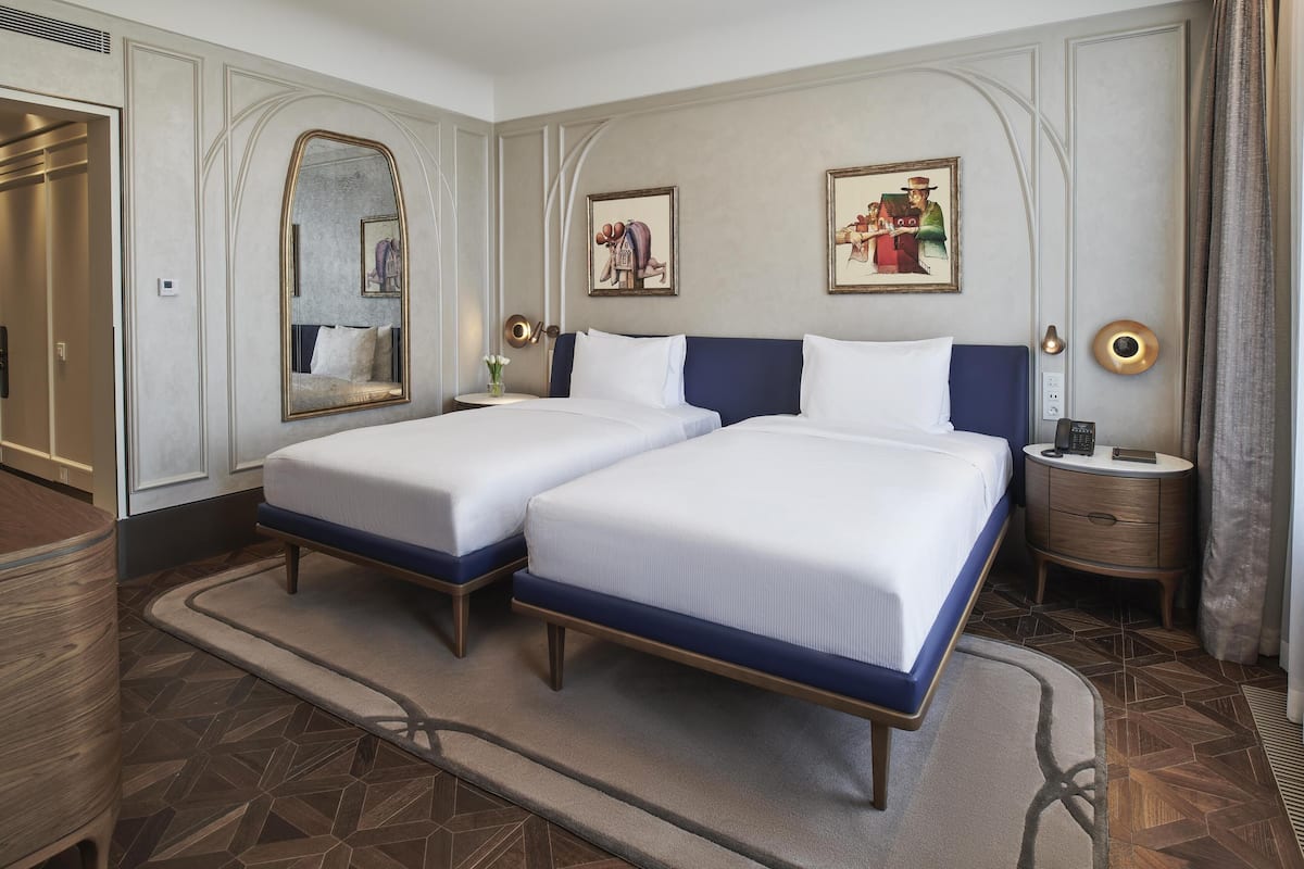 A hotel room with two beds, a mirror, a painting, a bedside table with a lamp, and a wardrobe.