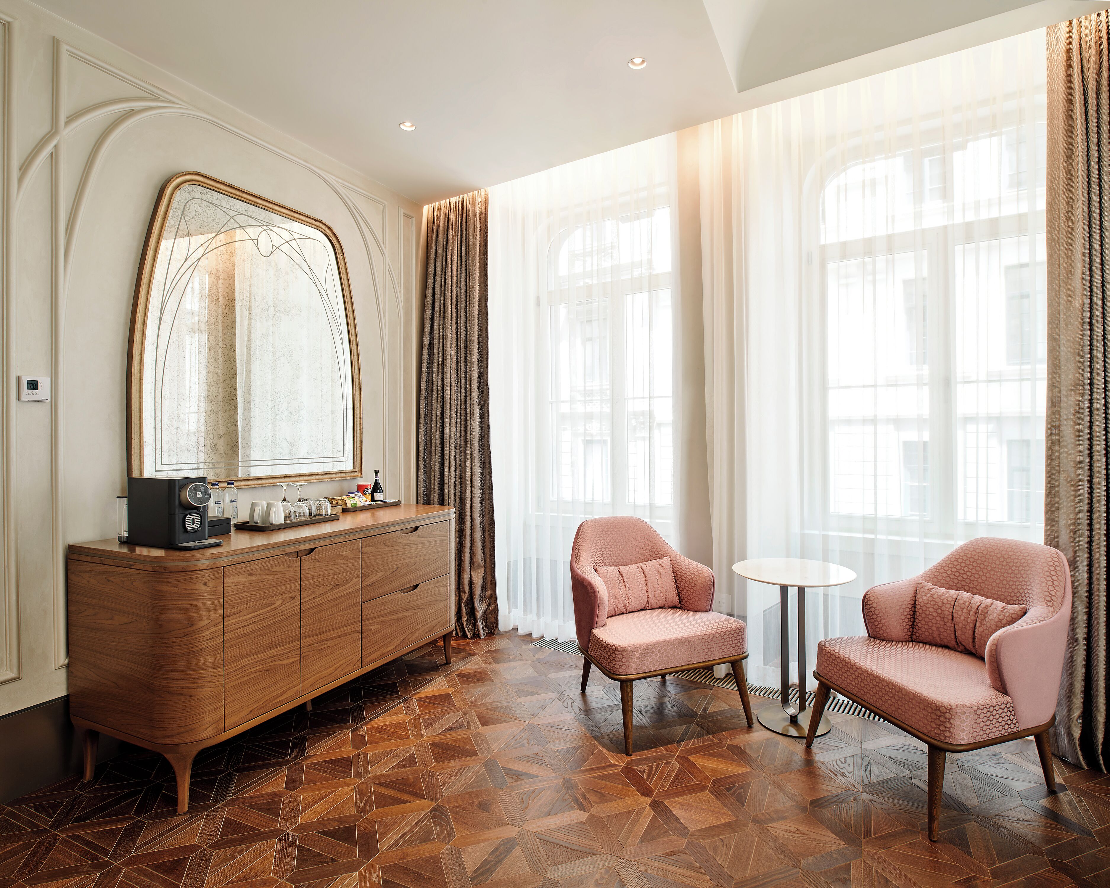 A hotel room with a wooden dresser, two pink armchairs, a round table, and large windows with sheer curtains.