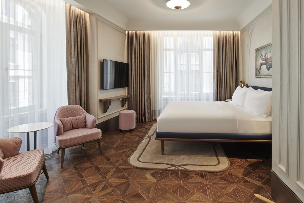 A hotel room with a bed, a TV mounted on the wall, a seating area with a chair and a small table, and a patterned floor.