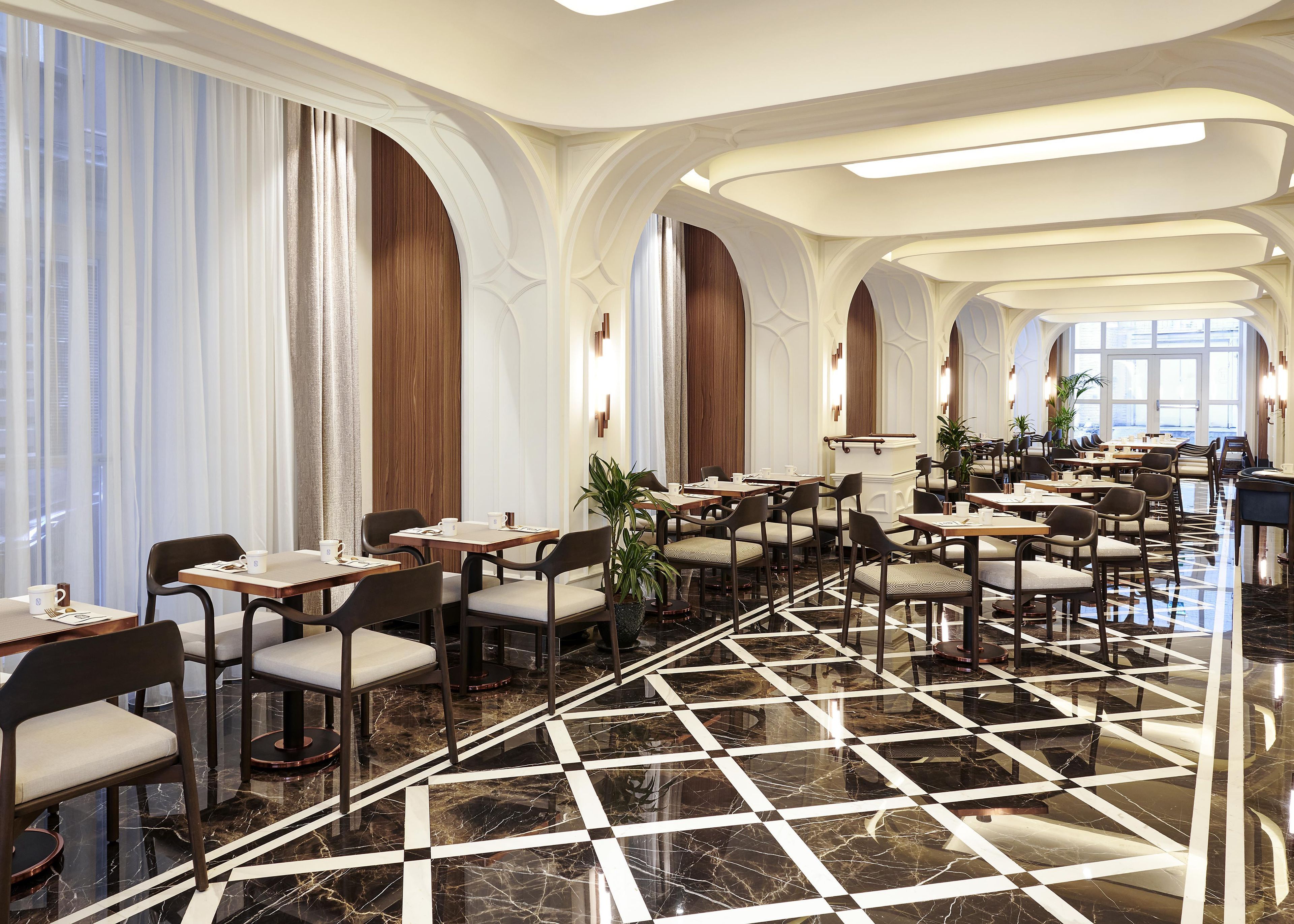 Ein geräumiger Hotel-Speisesaal mit mehreren Tischen und Stühlen, einem Schachbrett-Marmor-Boden und großen Fenstern mit Vorhängen.
