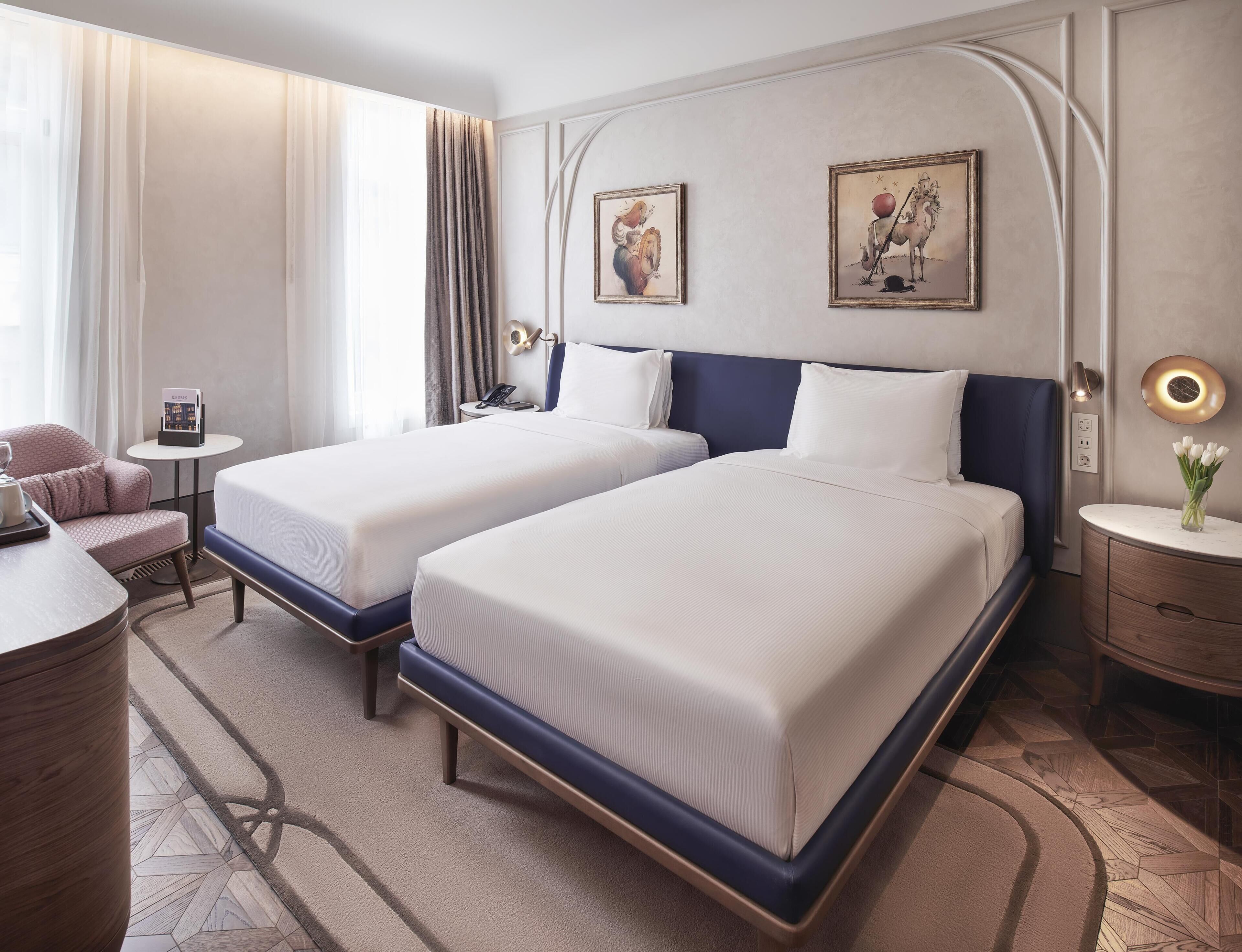 A hotel room with two beds, a desk, a chair, a nightstand, and a wall-mounted lamp.