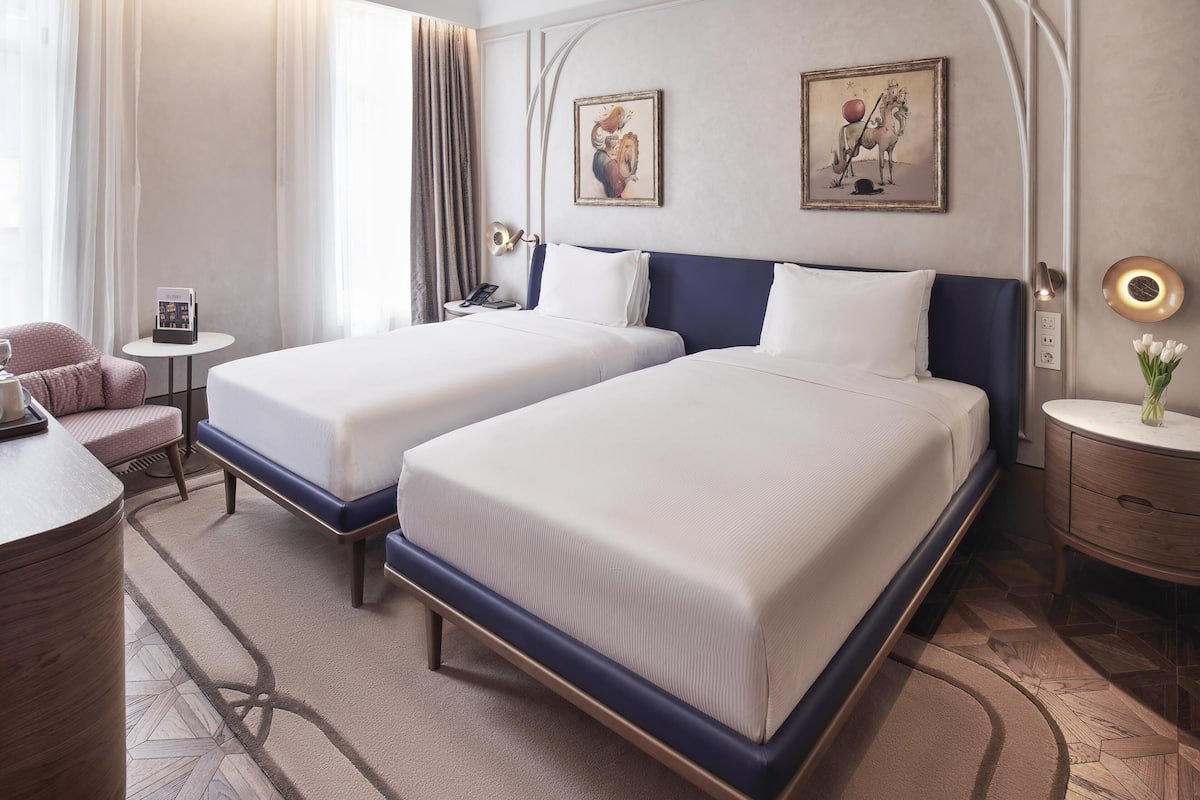 A hotel room with two beds, a desk, a chair, a nightstand, and a wall-mounted lamp.