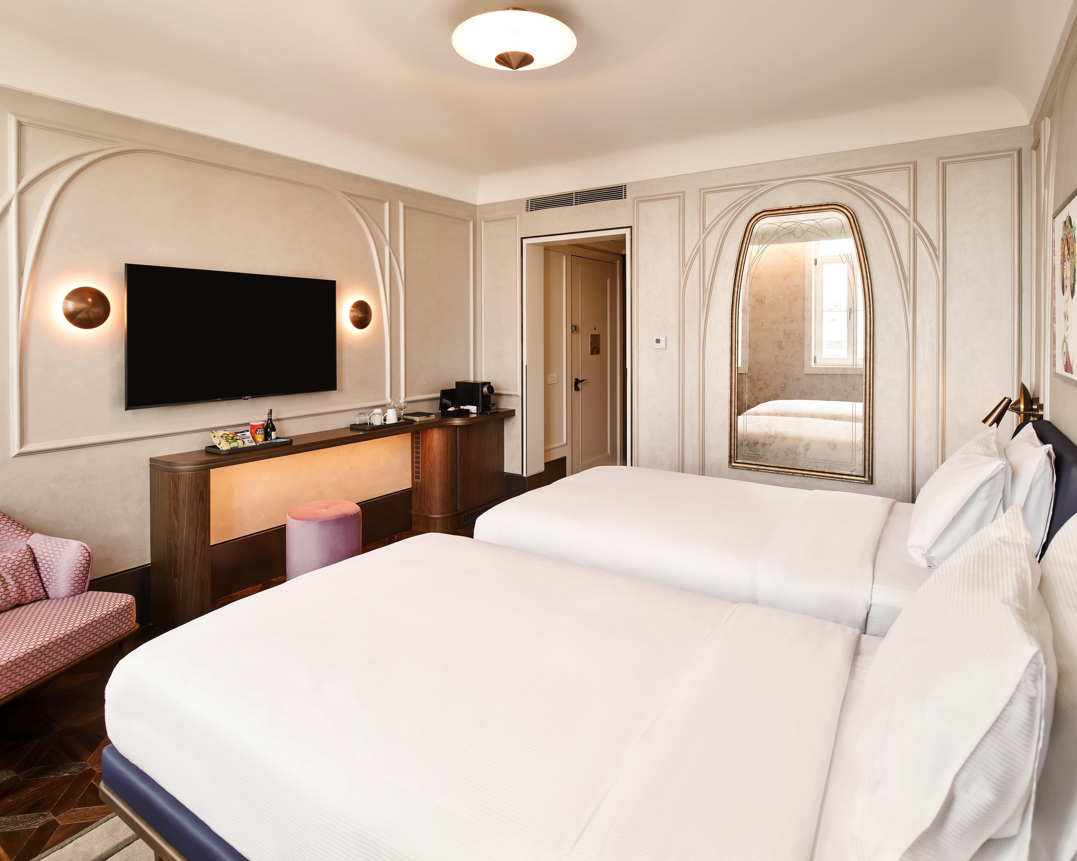 A hotel room with two beds, a TV mounted on the wall, a desk with a coffee machine, and a mirror with a decorative frame.