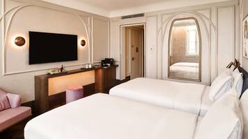 A hotel room with two beds, a TV mounted on the wall, a desk with a coffee machine, and a mirror with a decorative frame.