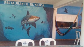 Interno di un ristorante con un murale raffigurante uno squalo e creature marine, un'amaca e un tavolo con tovaglia decorata.