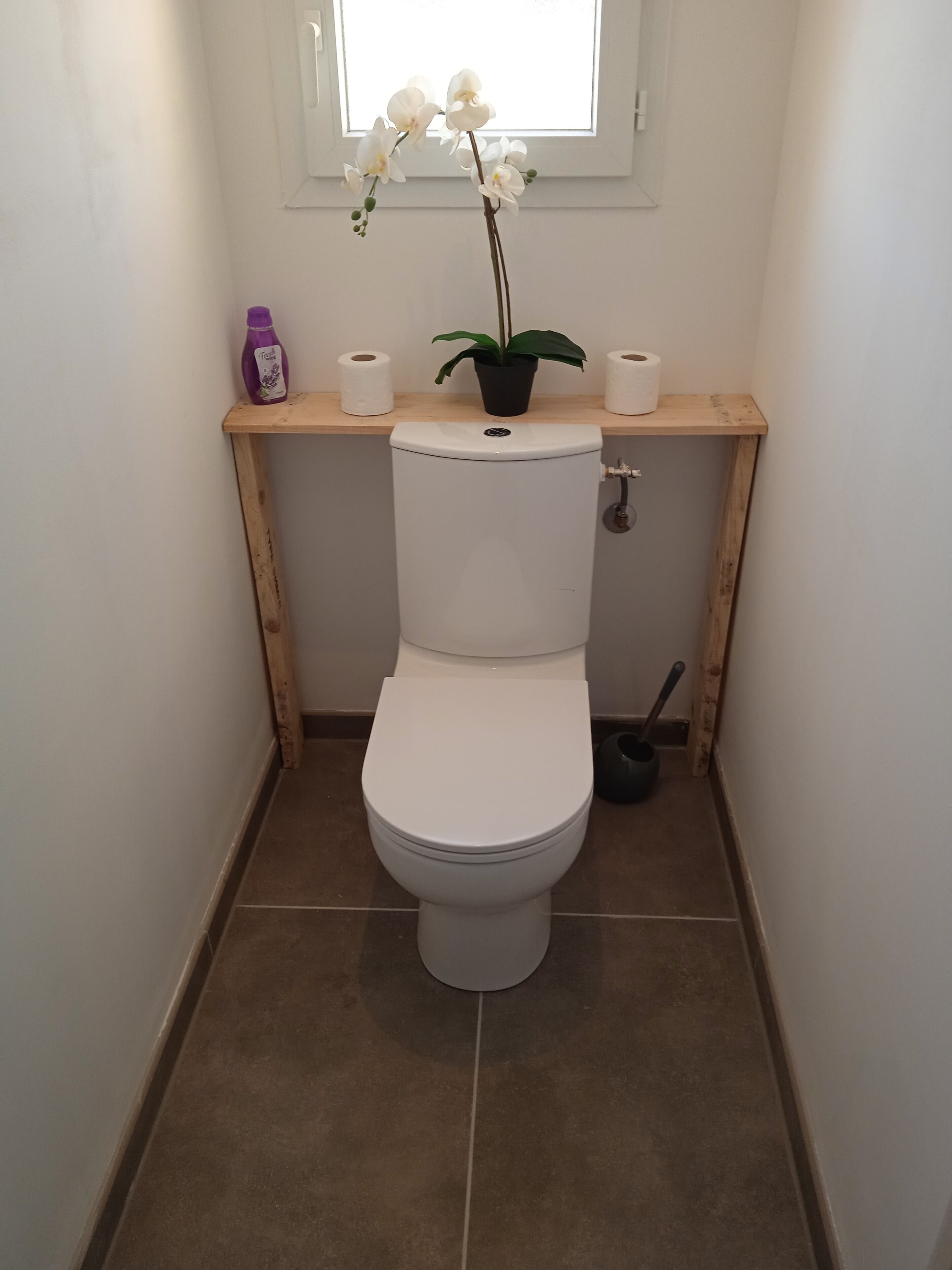 A small bathroom with a toilet, a shelf with toilet paper and a bottle, and a window above the toilet.