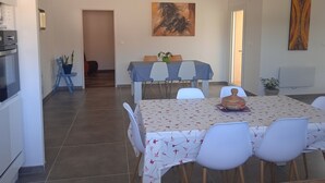 A dining area with a table set for four, surrounded by white chairs, a kitchen area with a stove, and a door leading to another room.