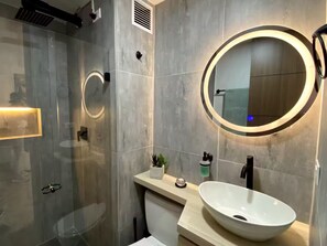 A modern bathroom with a glass shower enclosure, a round mirror with a light, and a white sink with a bowl.
